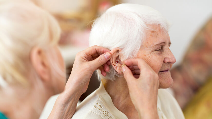 Elderly woman having a hearing aid fitted in her ear by another person indoors.