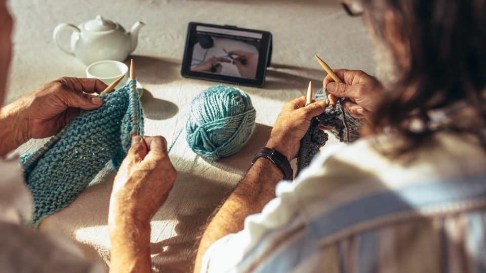 Two people knitting with teal yarn while watching a tutorial on a tablet, teapot and cup in the background.