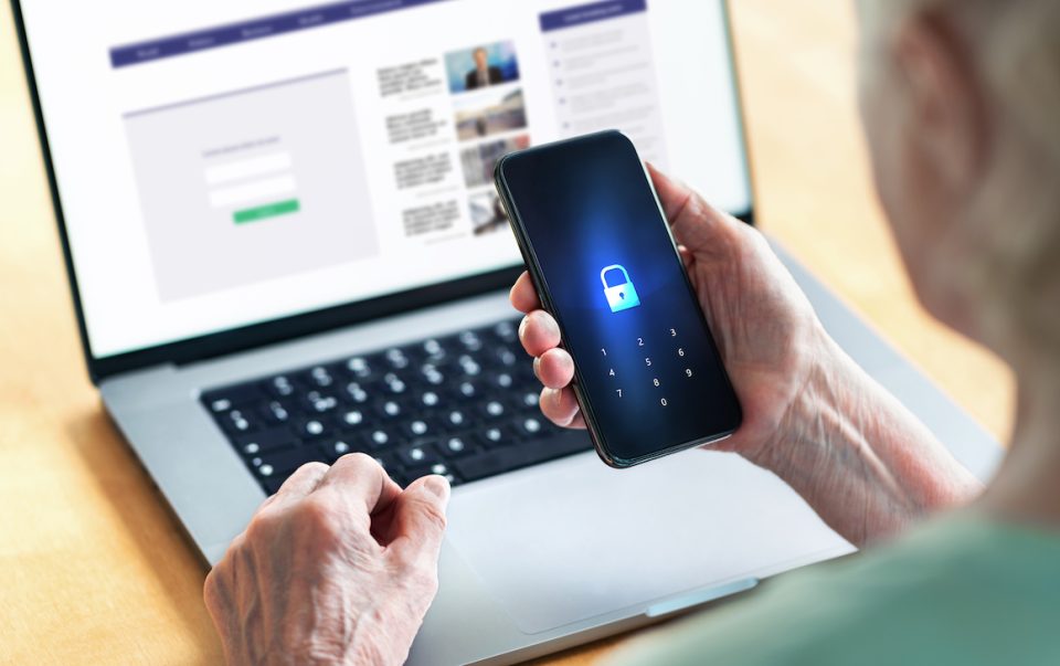 Person using a smartphone with a lock icon on screen, sitting in front of a laptop with a webpage open.