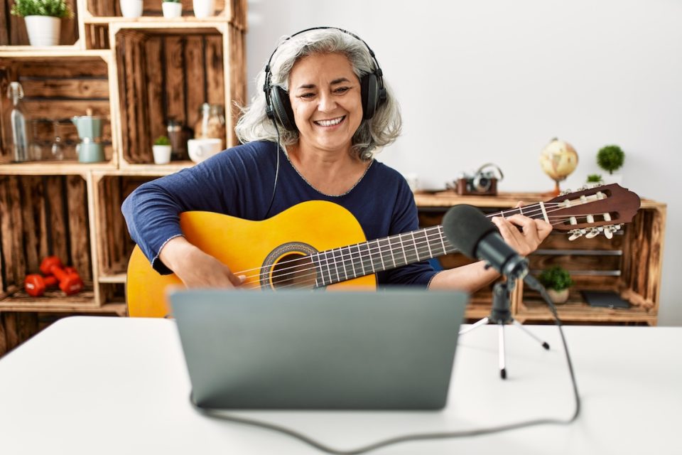 Woman with headphones playing guitar in front of a laptop and microphone, smiling while sitting at a white desk.