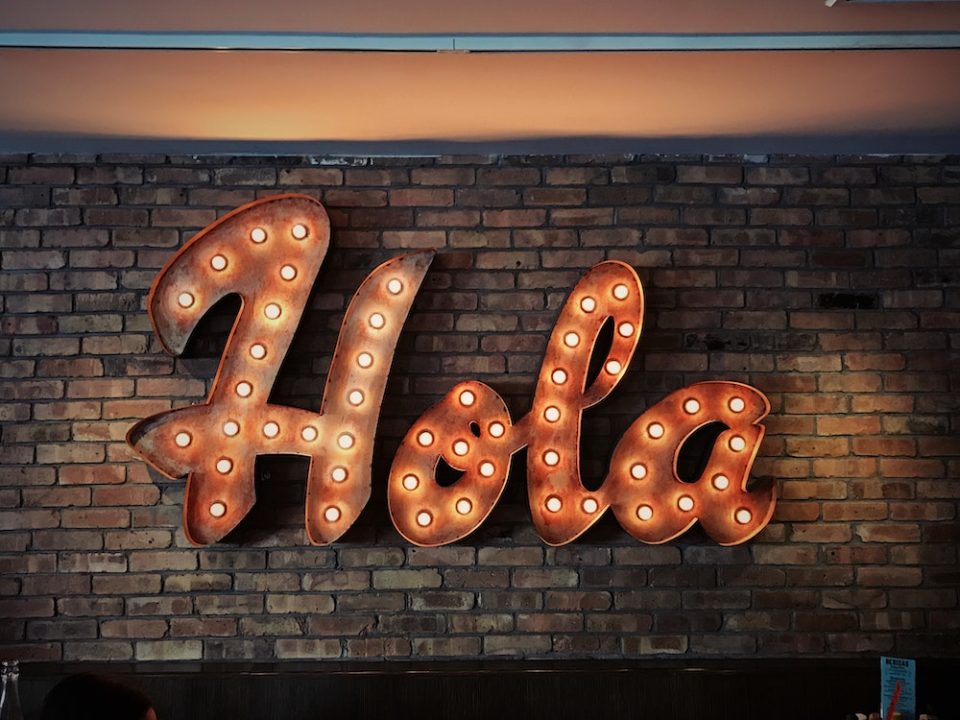 Illuminated "Hola" sign with warm lights mounted on a brick wall.